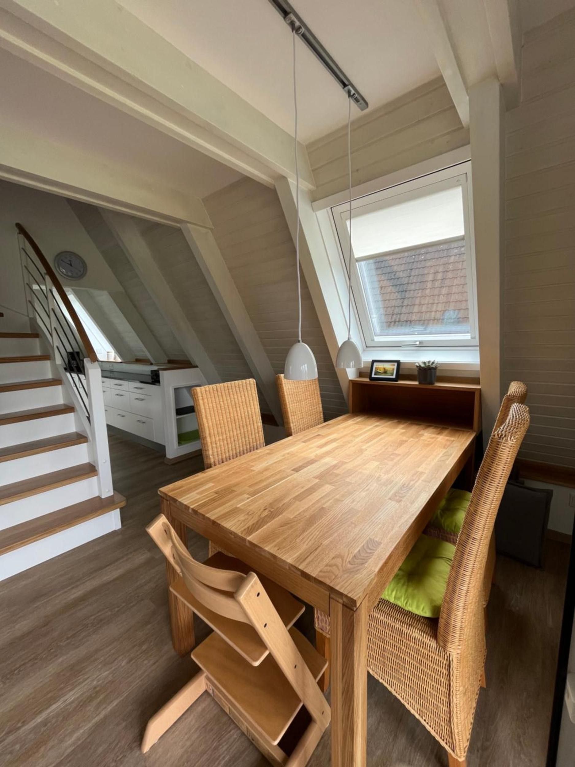 A dining area with a wooden table and wicker chairs, a window with blinds, and a staircase with white railings.