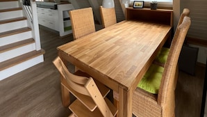 Zona pranzo con tavolo in legno e sedie in vimini, una finestra con persiane e una scala che conduce a un livello superiore.