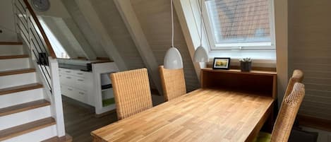 Un comedor con mesa de madera y sillas de mimbre, una ventana con persianas y una escalera con barandillas blancas.