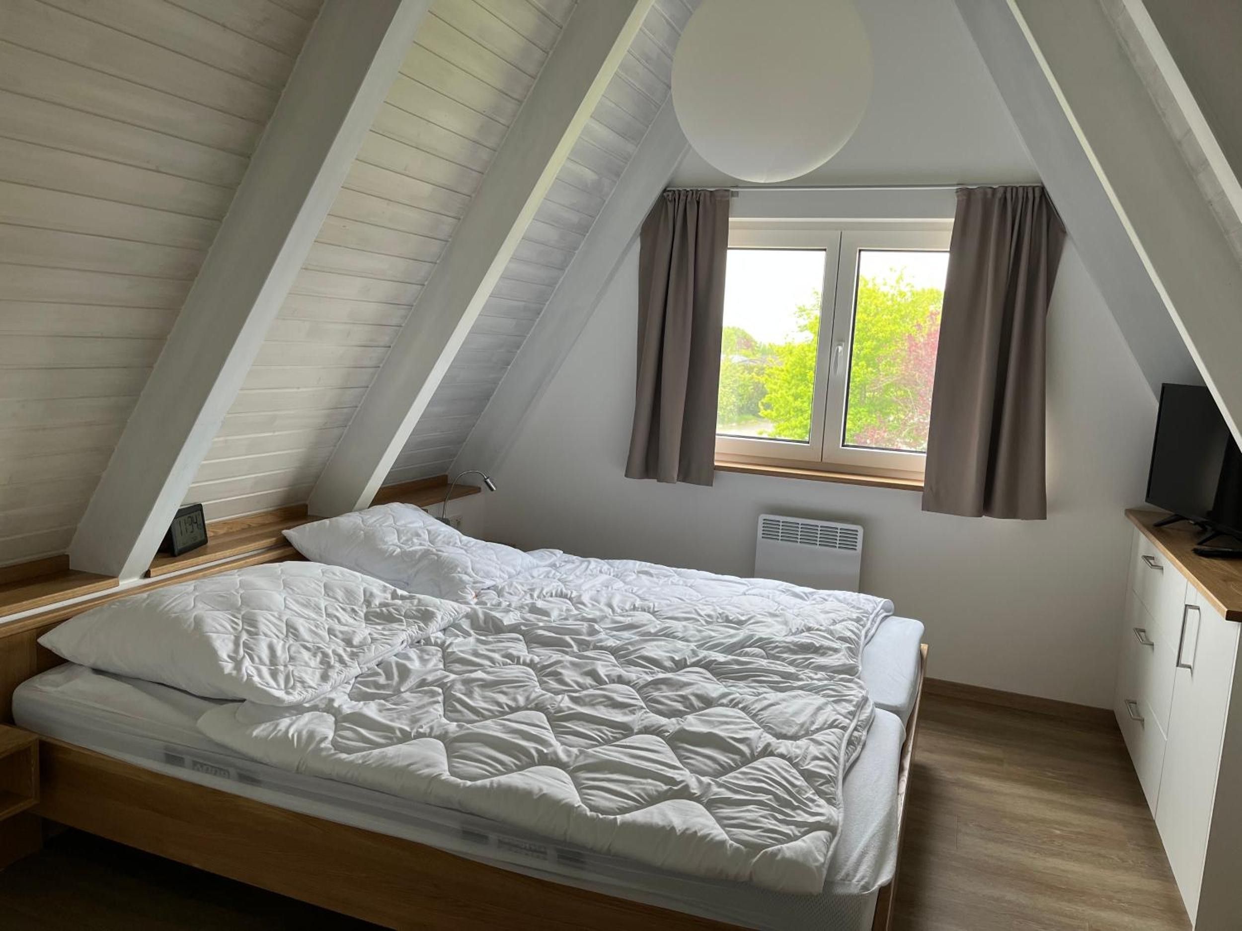 Ein Schlafzimmer mit schrägen Wänden, einem Bett, einem Fenster mit Vorhängen, einem Heizkörper und einem Fernseher auf einer Holzkabine.