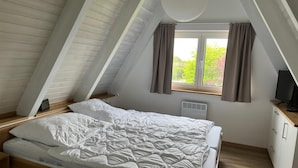 Una camera da letto con soffitto spiovente, un letto, una finestra con tende, un termosifone e una televisione su un mobile in legno.