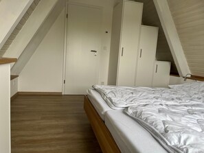 Ein Schlafzimmer mit schrägverdeckter Decke, einem Bett mit weißer Bettwäsche, Holzboden und weißen Kleiderschränken.