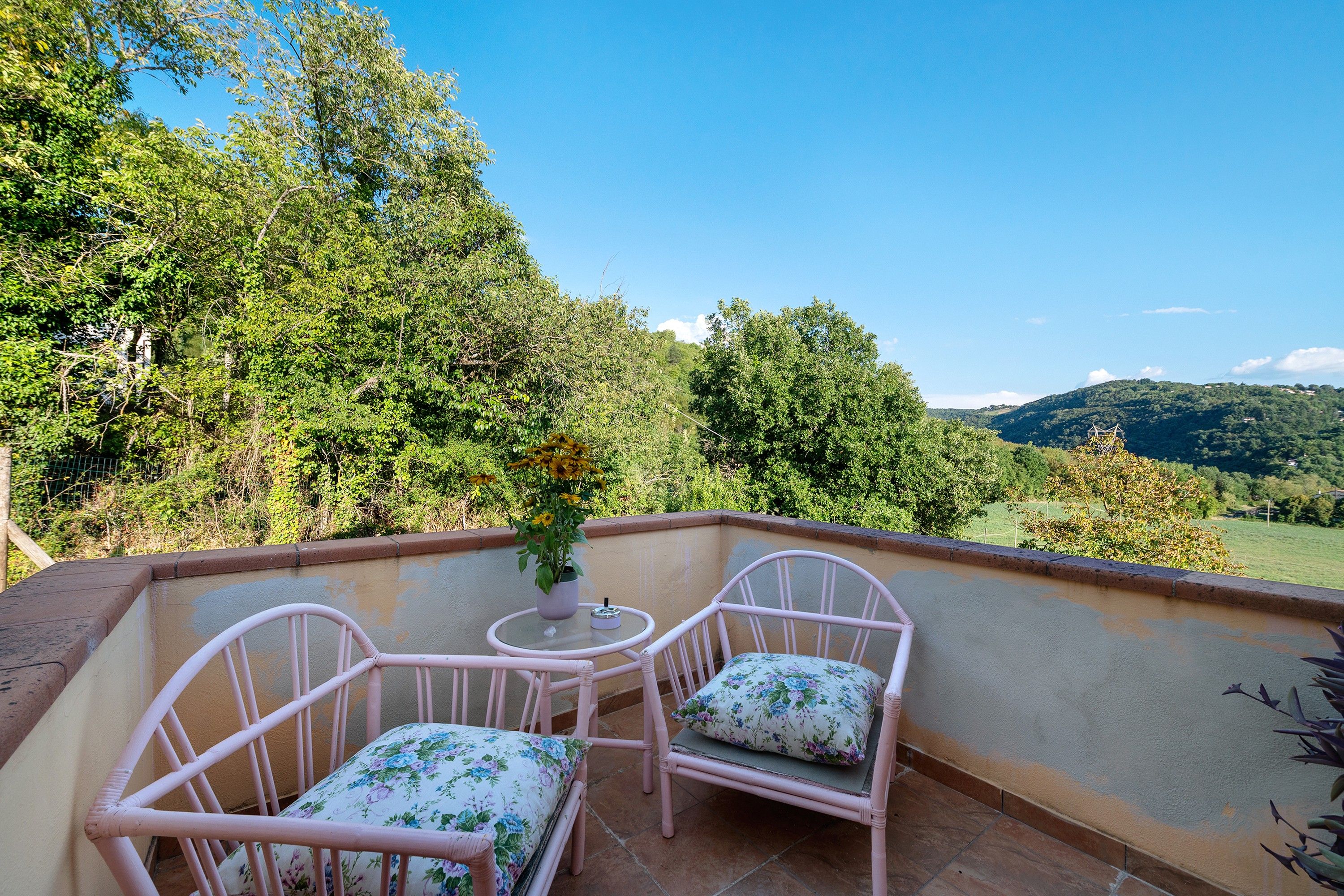 Ein Balkon mit zwei Korbstühlen und einem kleinen Tisch, mit Blick auf eine üppige, grüne Landschaft mit Hügeln.