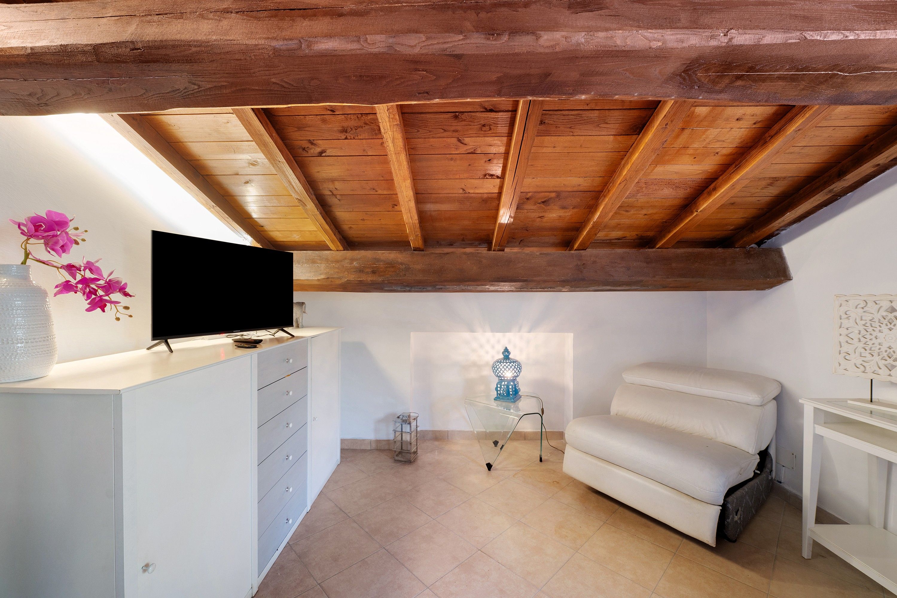 A room with a sloped ceiling, a flat-screen TV on a white dresser, a white sofa, and a small table with a blue lamp.