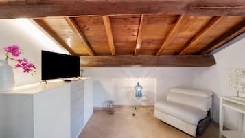 A room with a sloped ceiling, a flat-screen TV on a white dresser, a white sofa, and a small table with a blue lamp.