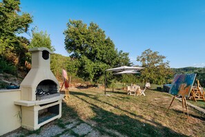 An outdoor setting with a stone-built barbecue area, a table with chairs, and an easel with a painting.