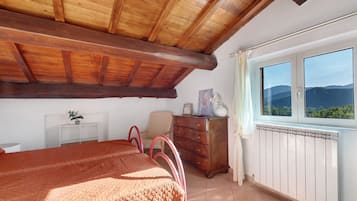 Une chambre avec un plafond en bois, un lit recouvert d’une couverture rouge, une commode, une chaise, une fenêtre donnant sur la montagne et un radiateur.