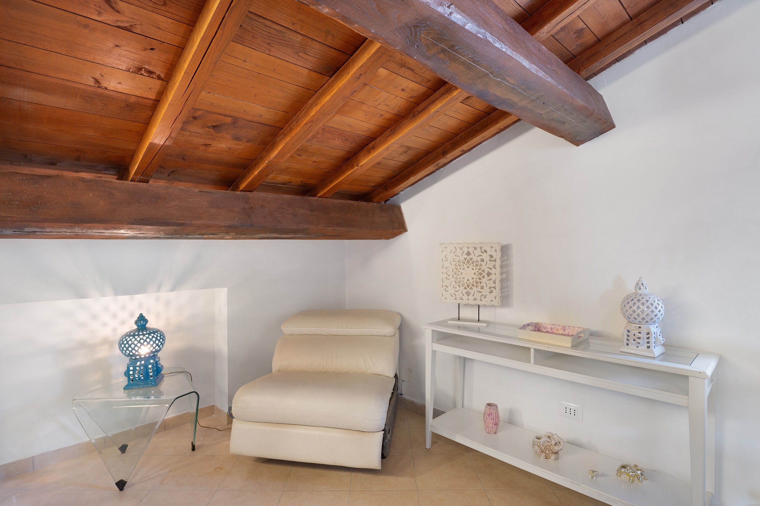 Een kamer met een schuine wand, een witte fauteuil, een glazen bijzettafeltje, een witte console met decoratieve objecten, en een wandlamp.