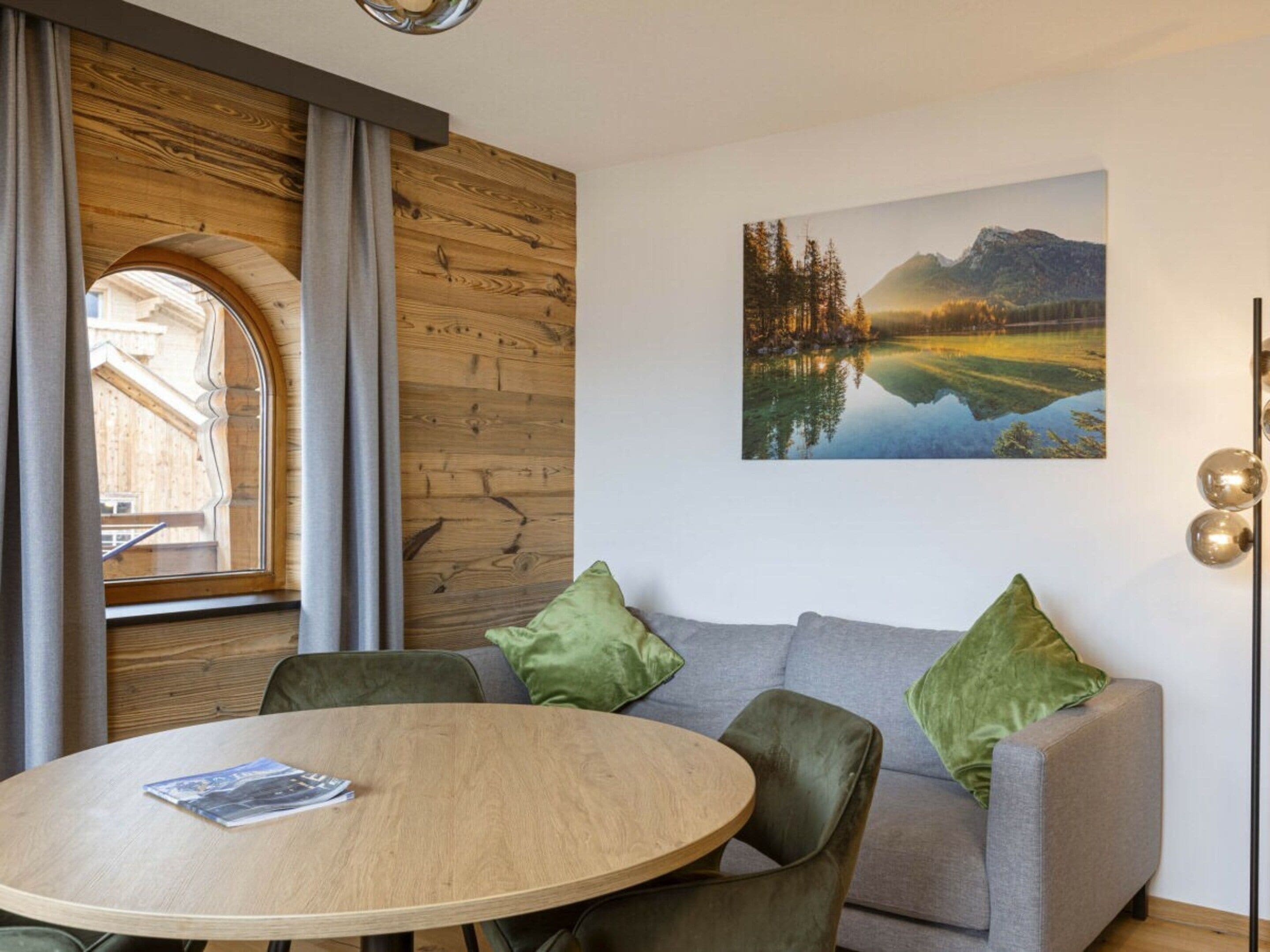A room with a round wooden table, a grey sofa with green cushions, a wooden wall panel, and a framed landscape painting.