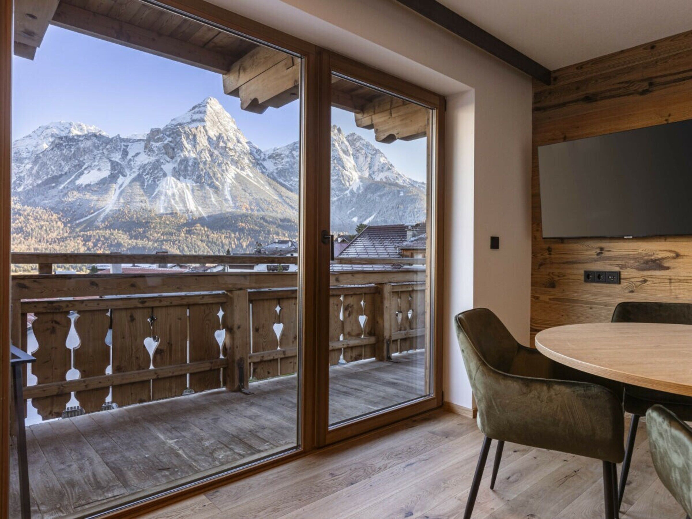 A room with a wooden floor, a dining table with chairs, a TV mounted on the wall, and a large sliding glass door leading to a balcony with a mountain view.