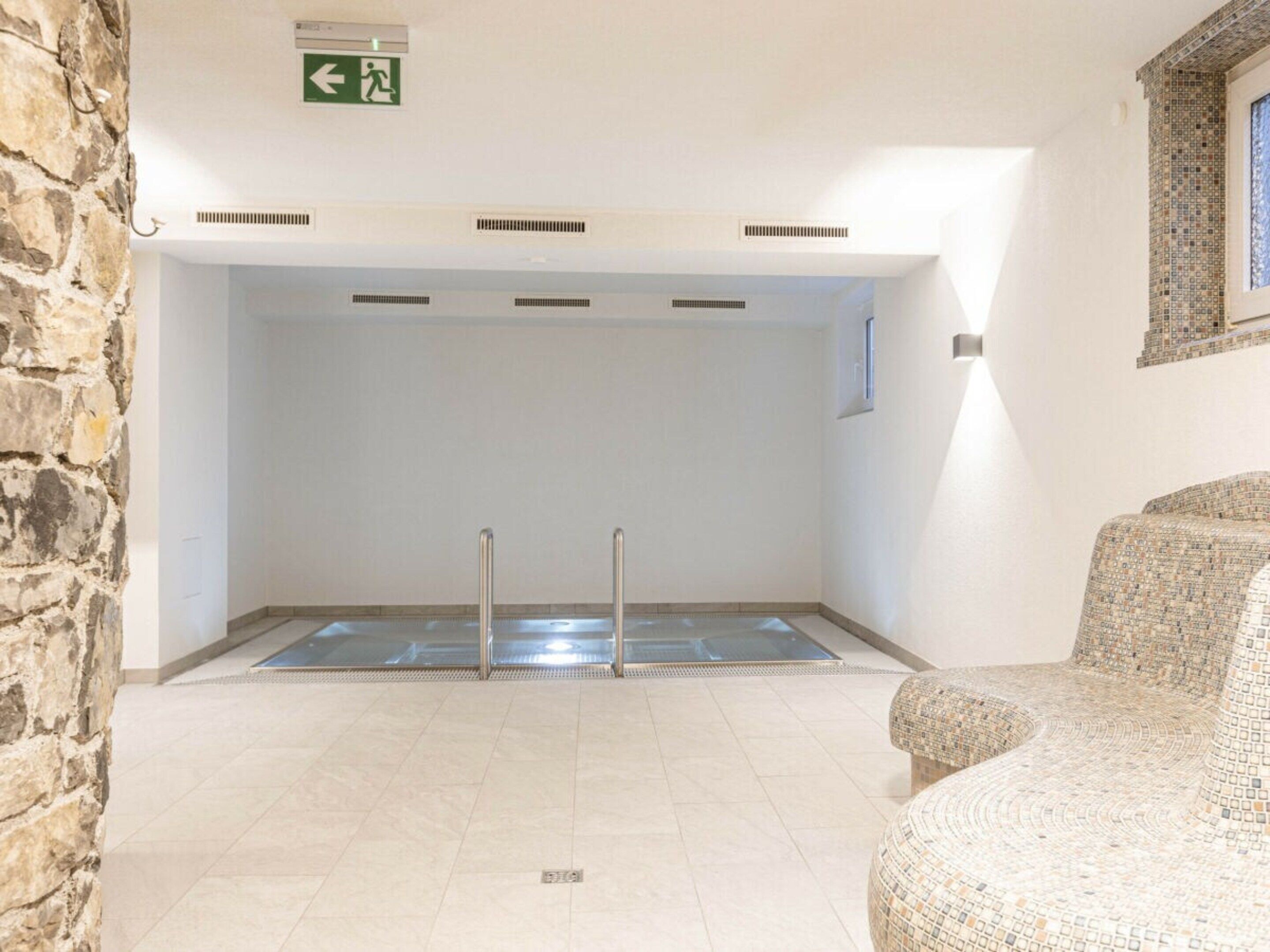 A room with a pool, stone wall, and a green exit sign.