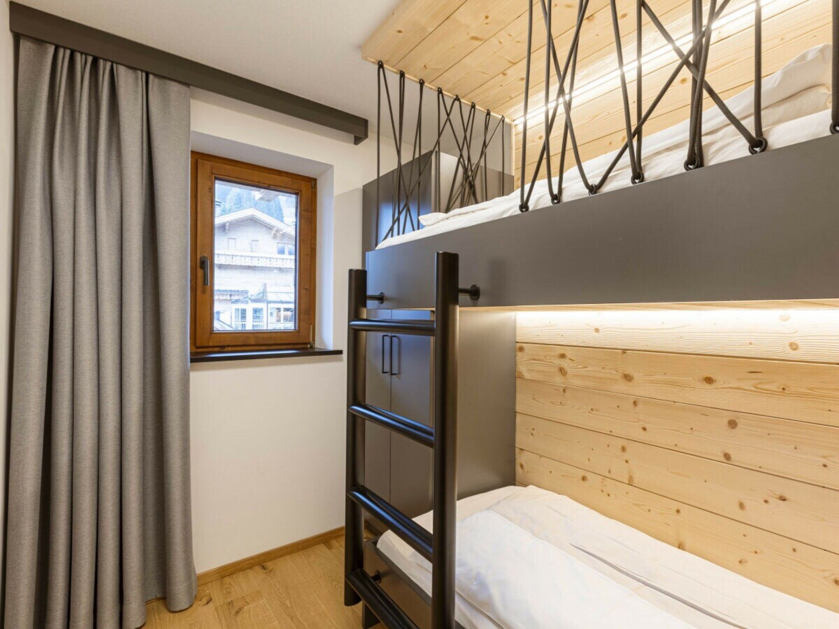 A compact room with a bunk bed, a window with curtains, a wooden ceiling, and a metal ladder.