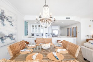 A dining area with a chandelier, woven chairs, and a central table with placemats. - Updated Luxury Gulf-Front 3BR | Beach Club 1404, Pools, Spa & Beach Chairs (Pensacola Beach)