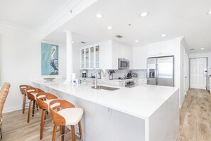 A modern kitchen with white cabinetry, stainless steel appliances, and a central island with bar stools. - Updated Luxury Gulf-Front 3BR | Beach Club 1404, Pools, Spa & Beach Chairs (Pensacola Beach)