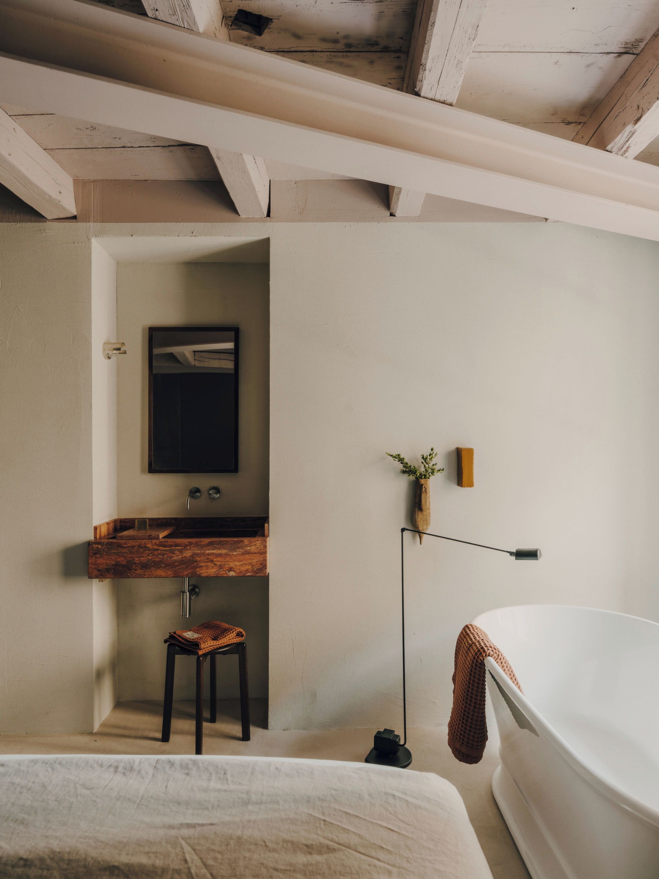 A bedroom with a large bed, a wooden vanity, a small stool, a wall-mounted light, and a bathtub.