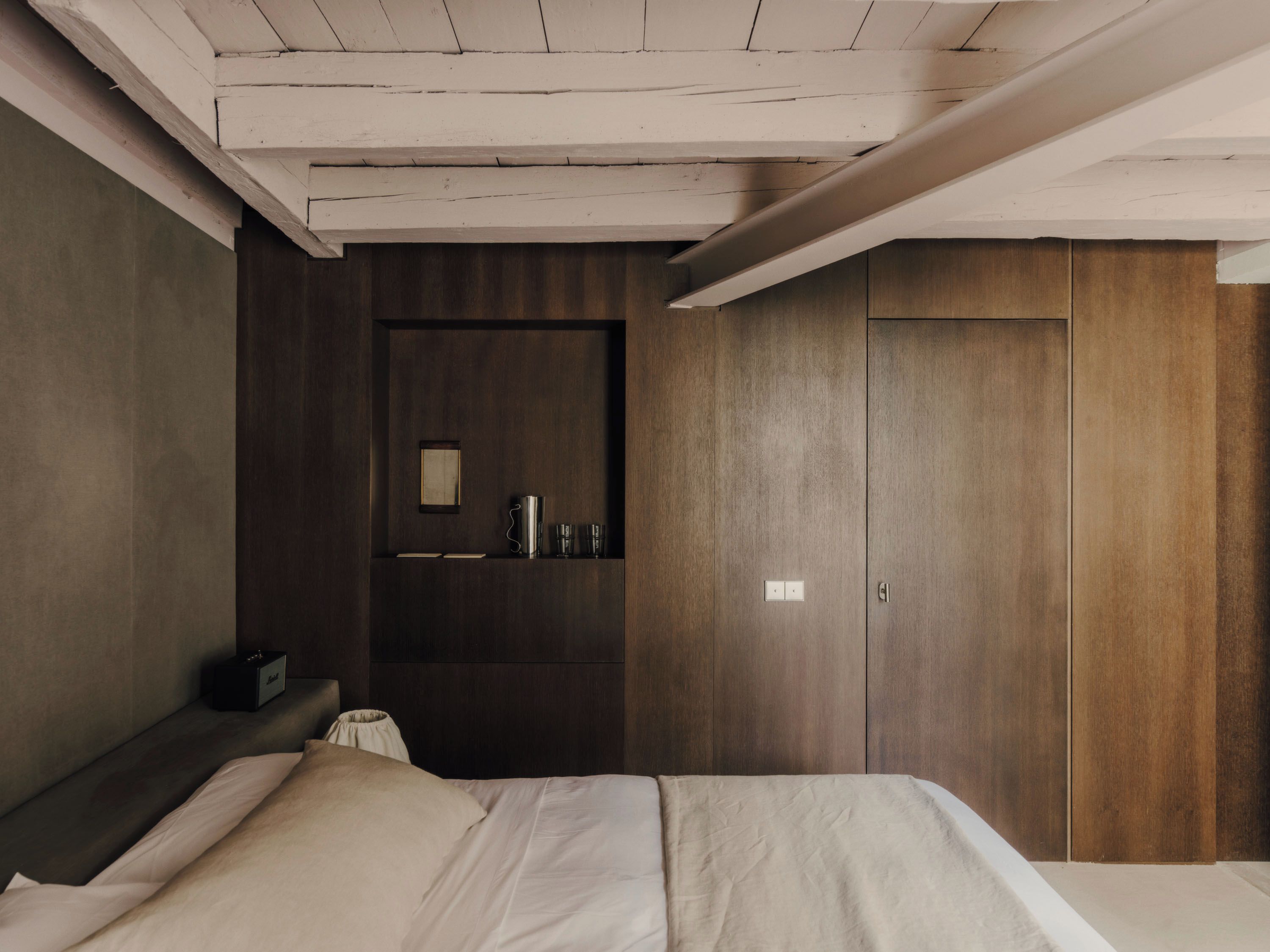 A bedroom with a bed, a wardrobe, a shelf with items, and a light switch.