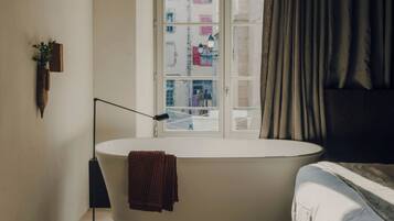 A bathroom with a bathtub, a window with curtains, a floor lamp, and a towel hanging on the wall.