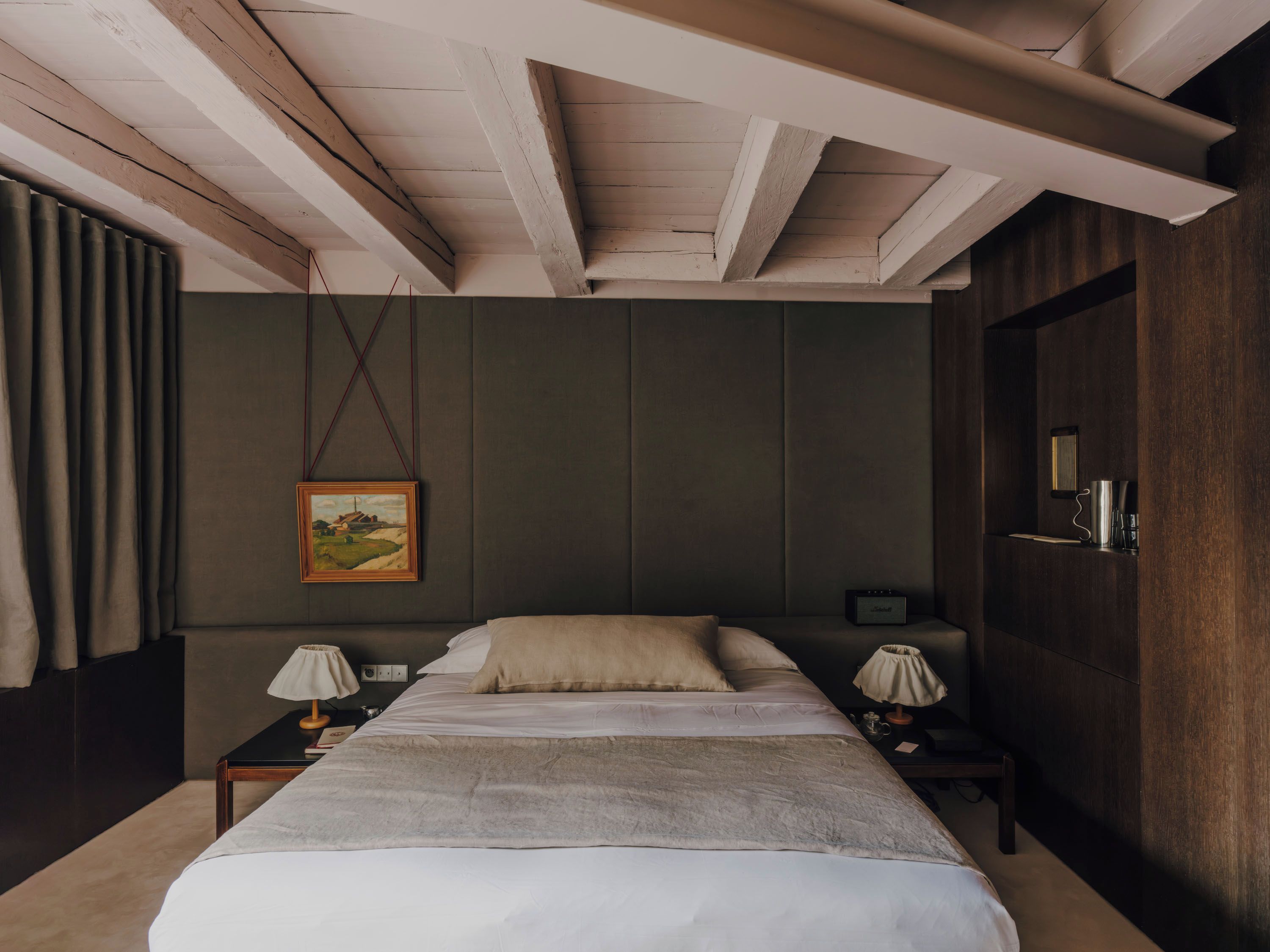 A bedroom with a bed, two bedside tables, a lamp, a painting, and a nightstand with a radio.