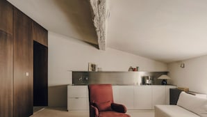 Una habitación de hotel moderna con un sillón rojo, un sofá blanco, una mesita de café pequeña y una kitchenette al fondo.
