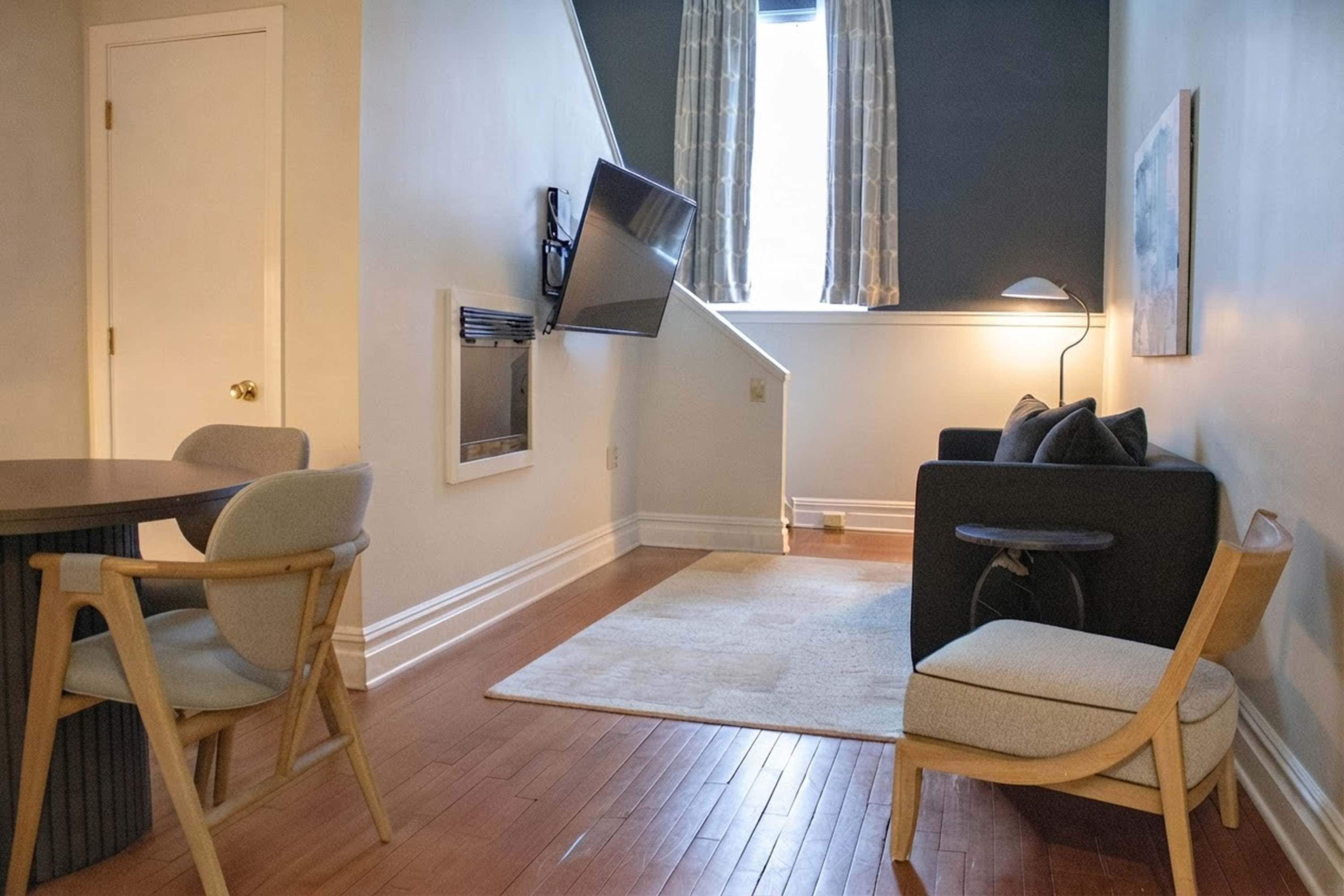 Une pièce comprenant une table à manger, deux chaises, un canapé, un téléviseur fixé au mur, une lampe et une fenêtre avec des rideaux.