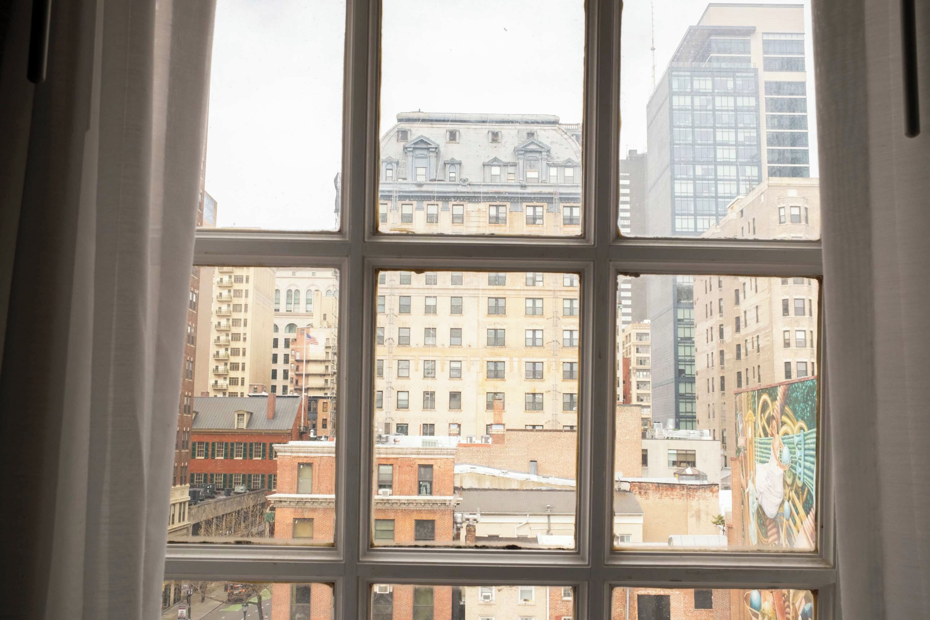A view through a window with a grid pattern, displaying a cityscape with various buildings, including a prominent brick building and a modern high-rise.