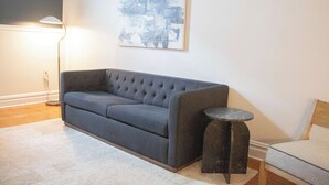 Ein Wohnzimmer mit grauem Sofa, einem kleinen runden Tisch, einer Stehlampe und einem abstrakten Kunstwerk an der Wand.