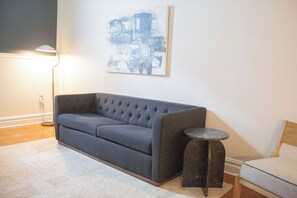 Ein Wohnzimmer mit grauem Sofa, einem kleinen runden Tisch, einer Stehlampe und einem abstrakten Kunstwerk an der Wand.