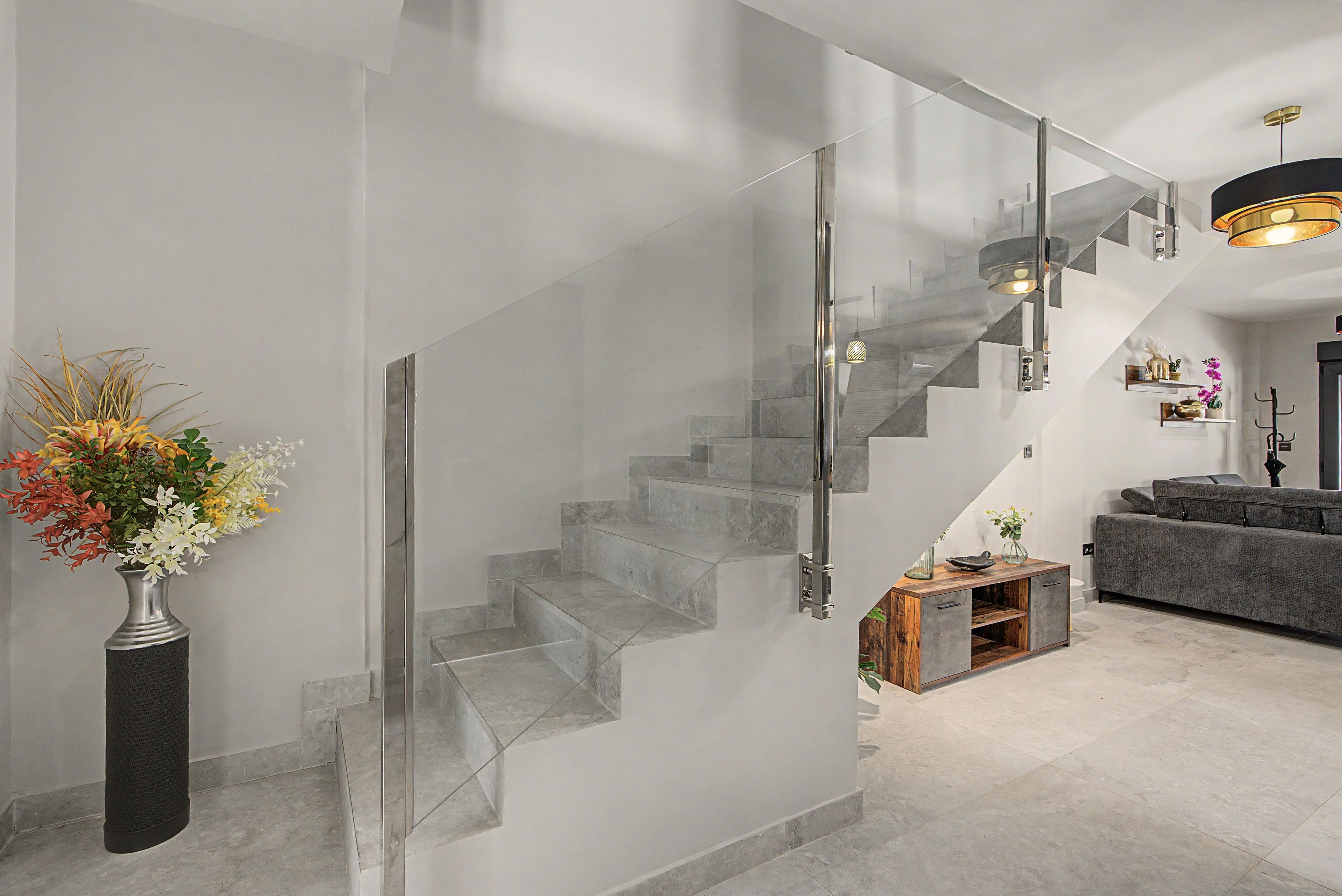 Un intérieur moderne avec un escalier en verre, un vase rempli de fleurs et un canapé gris.