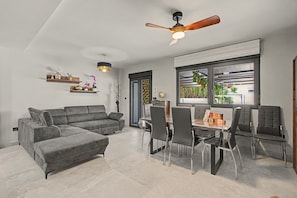 A modern living room with a grey sofa, a dining table with chairs, a ceiling fan, and a window with a view of greenery.