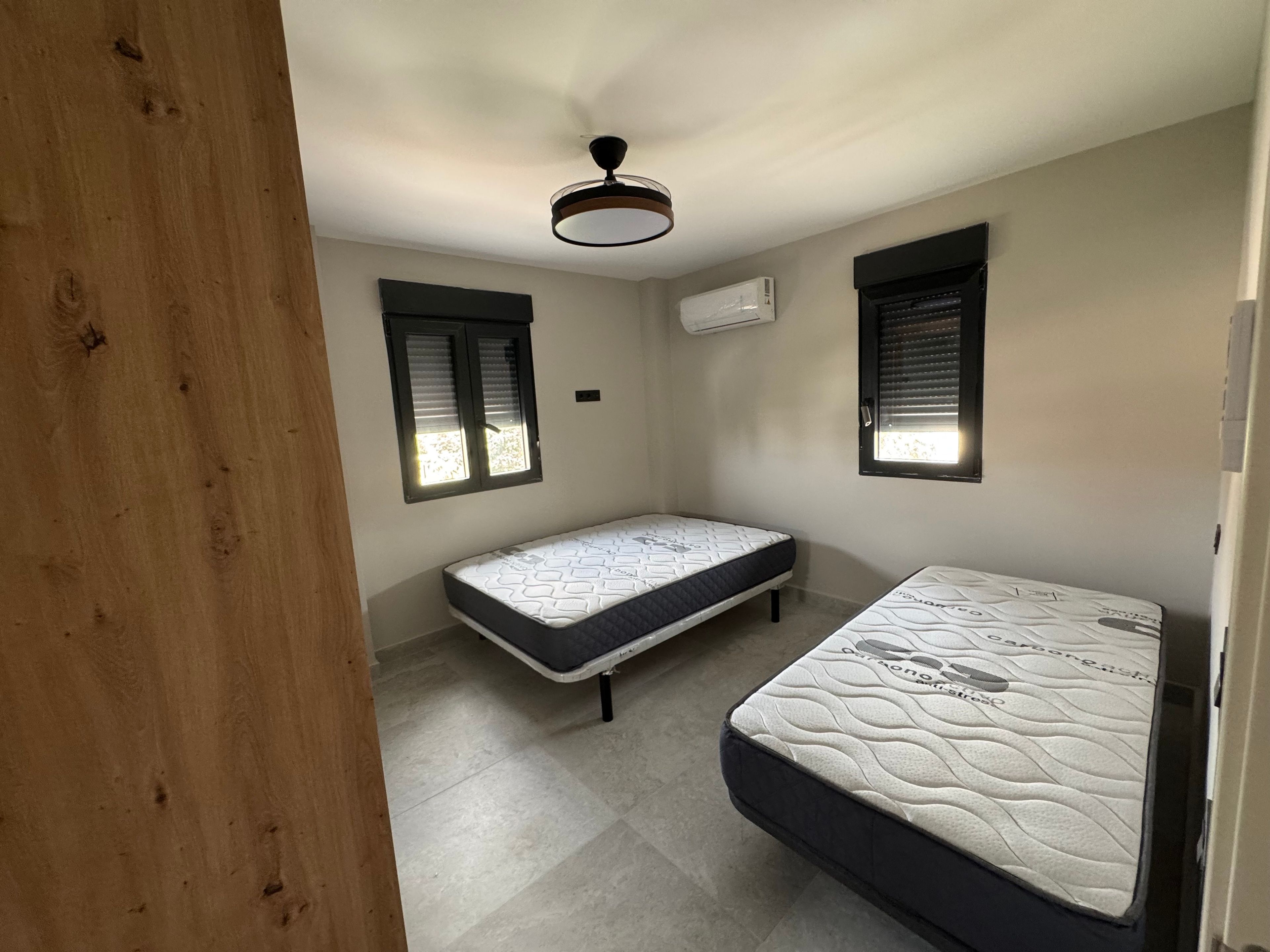 A room with two beds, a ceiling fan, and two windows with blinds.