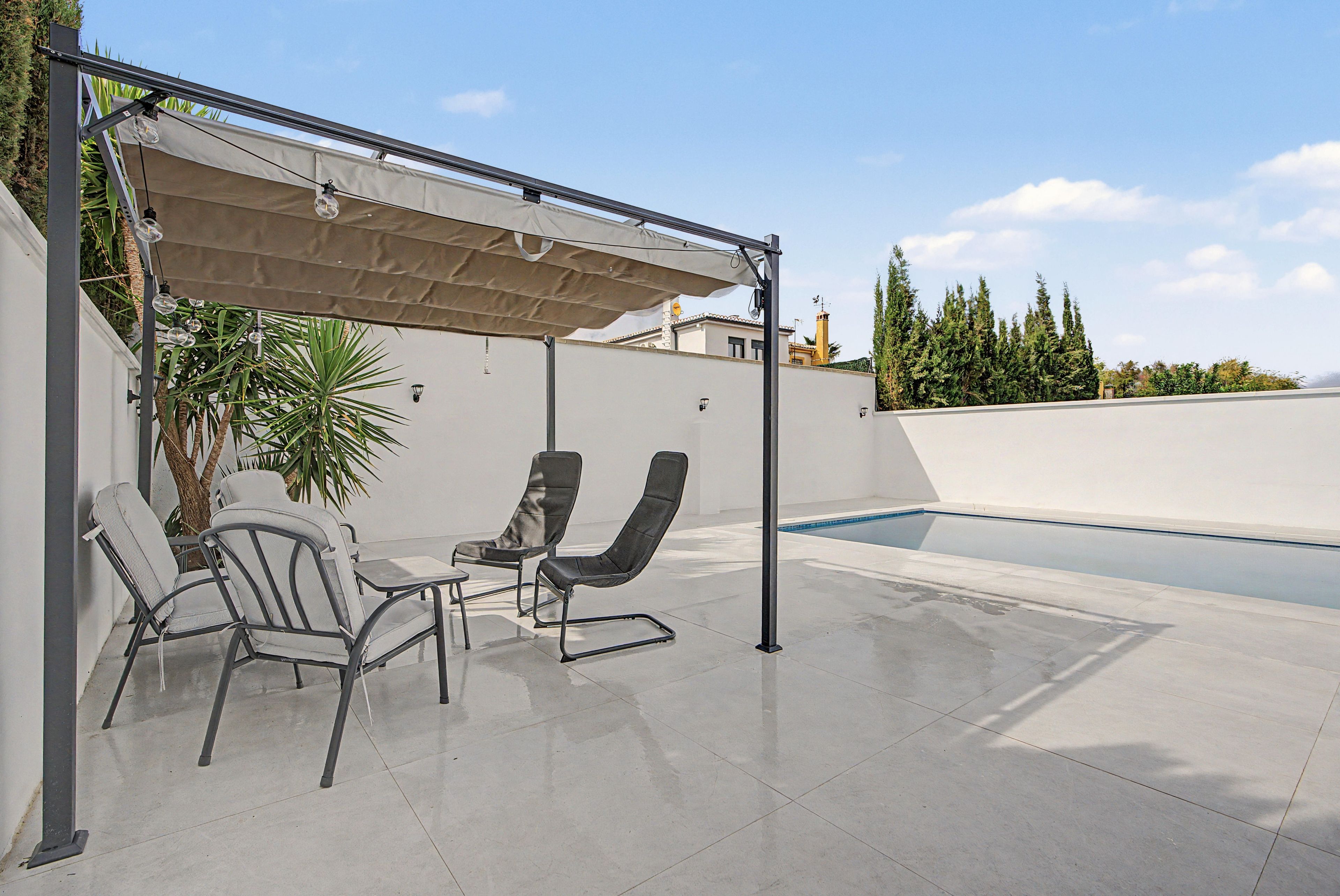 Un espace extérieur abrité avec deux chaises et une table, une piscine et une vue sur un bâtiment et des arbres en arrière-plan.