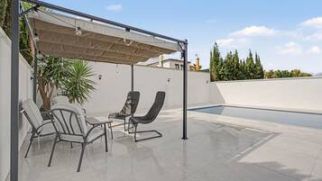 Un espace extérieur abrité avec deux chaises et une table, une piscine et une vue sur un bâtiment et des arbres en arrière-plan.