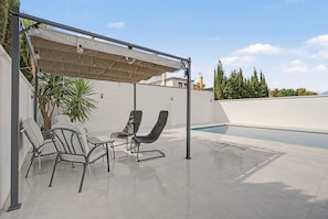 A covered outdoor seating area with two chairs and a table, a swimming pool, and a view of a building and trees in the background.