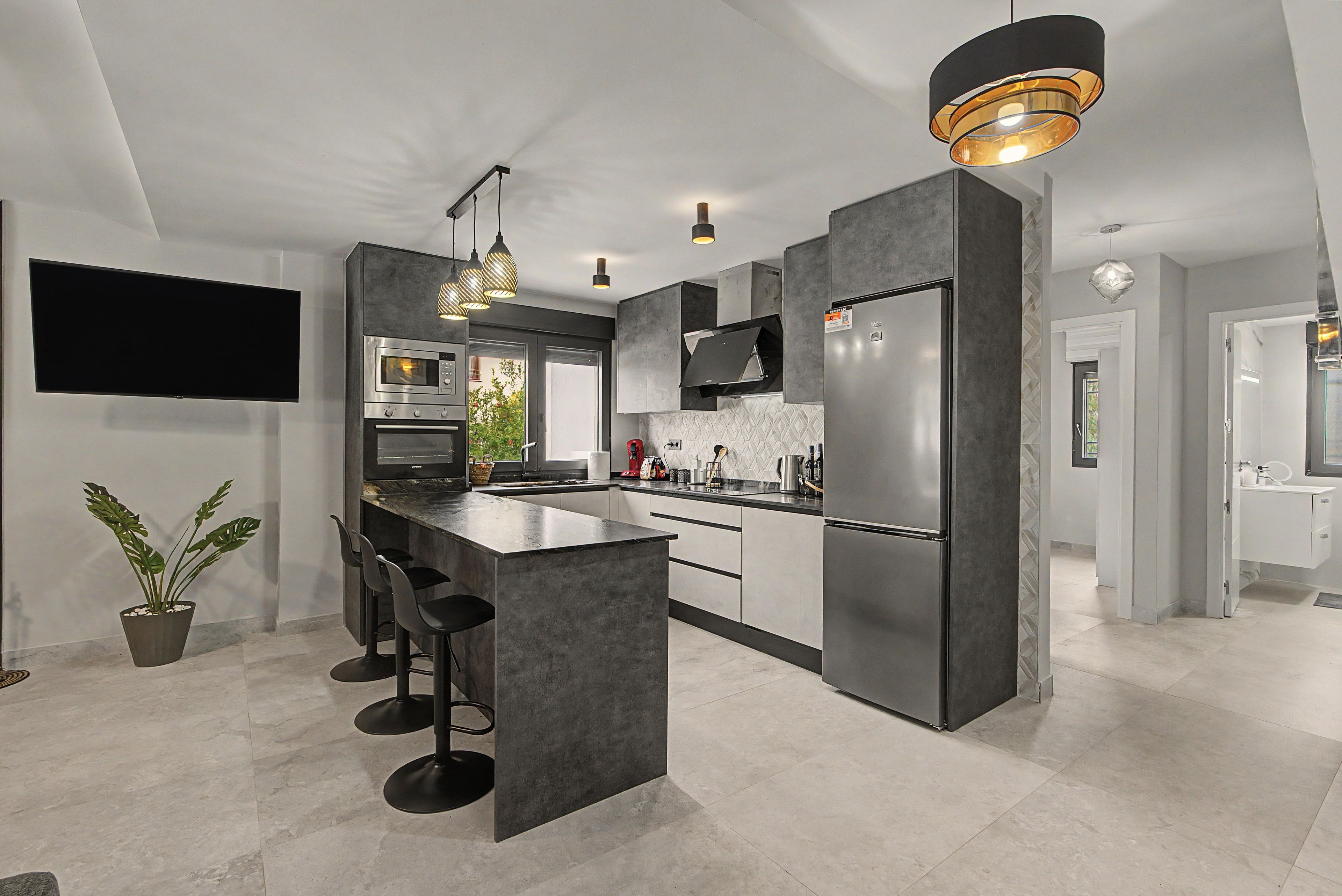 Een moderne keuken met een centraal eiland, roestvrijstalen apparatuur, hanglampen en een flatscreen televisie aan de muur.