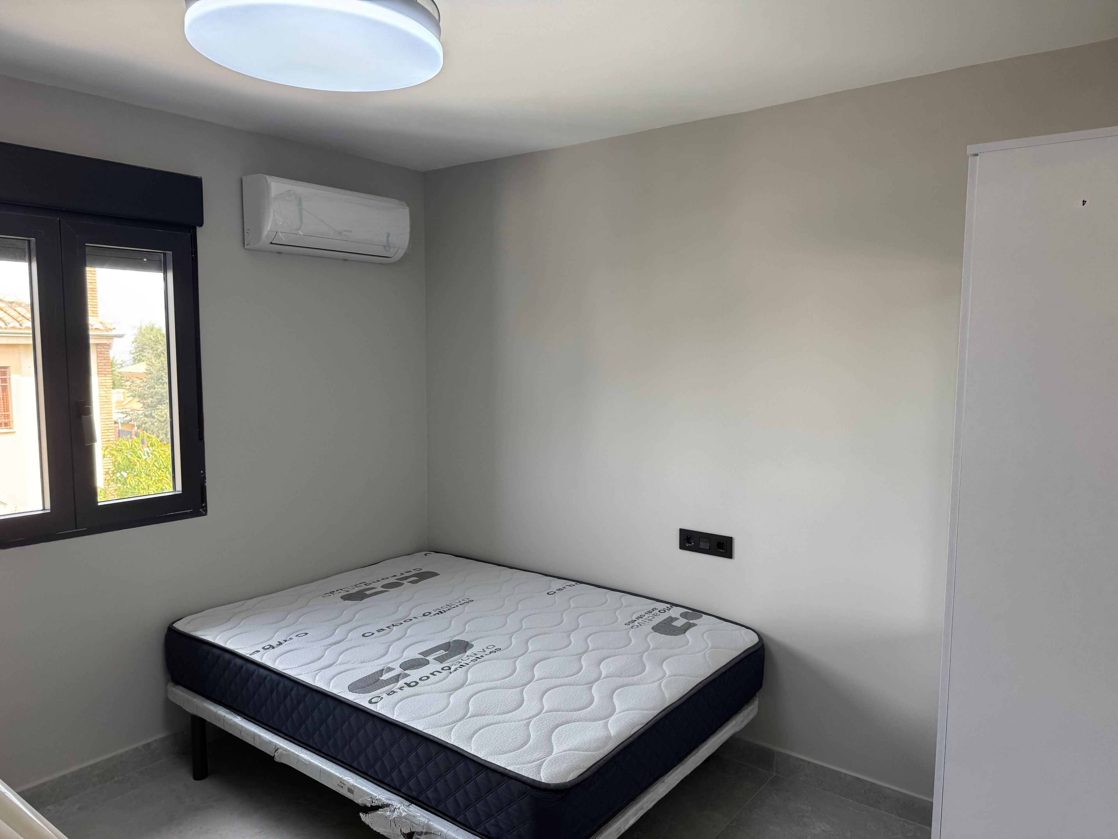 A small, clean room with a single bed, a window, and an air conditioning unit.