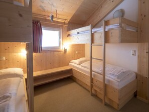 Habitació amb literes, parets de fusta, una finestra amb cortines i una petita estant.