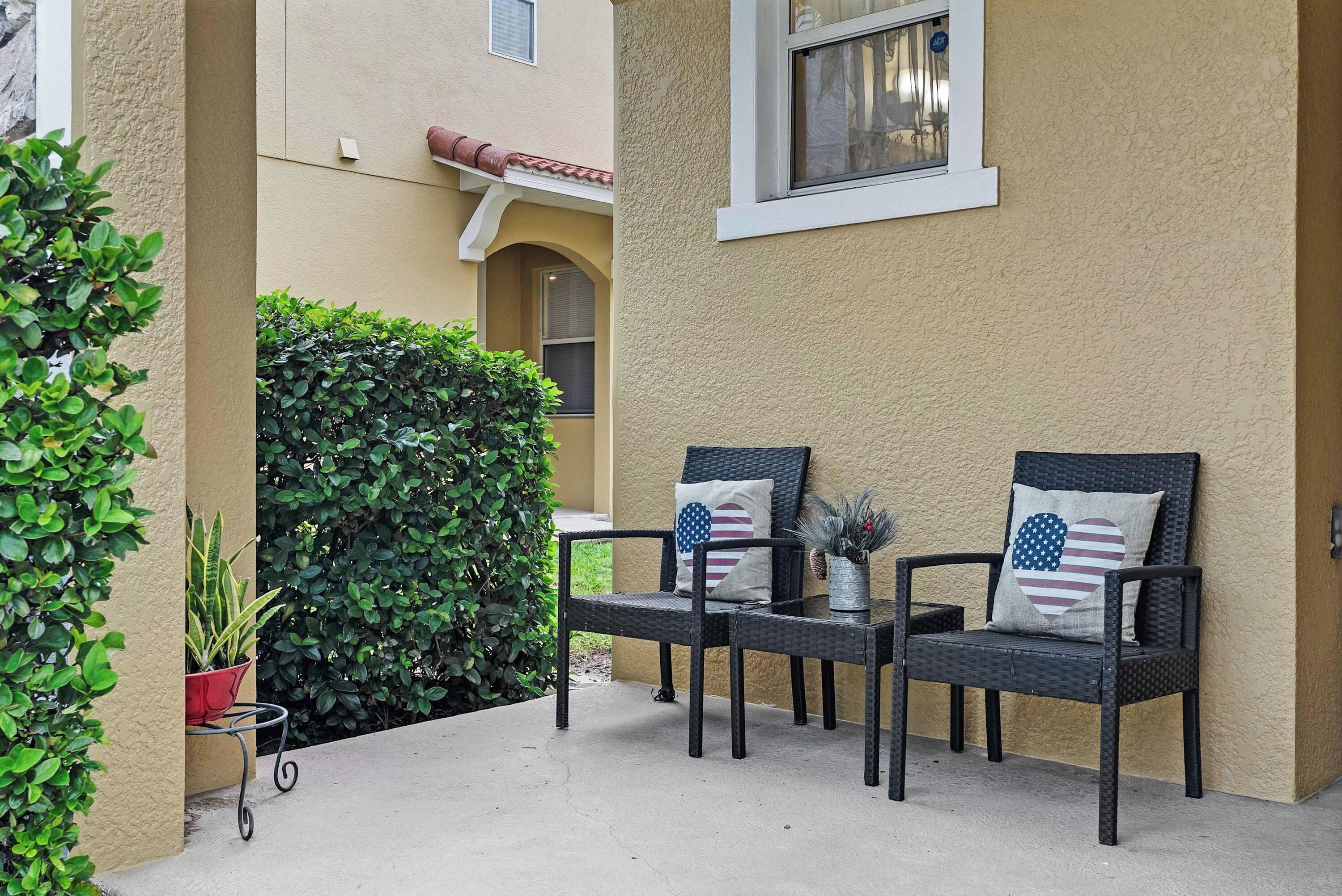 An outdoor seating area with two chairs and a small table, surrounded by greenery and a building facade.