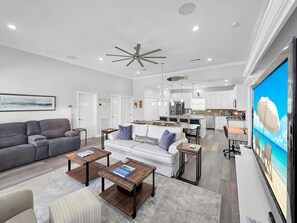 A spacious living room with a grey sofa, a white sofa, and a coffee table. There is a kitchen in the background with white cabinets and a large window. - Mer Sea 4 Bedroom Home (Port Bolivar)