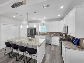 A modern kitchen with white cabinetry, a granite island with bar stools, stainless steel appliances, and pendant lighting. - Mer Sea 4 Bedroom Home (Port Bolivar)