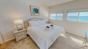 Ein Schlafzimmer mit einem großen Bett, zwei Nachttischen, einem Beistelltisch, einem gerahmten Bild und Meerblick durch die Fenster.