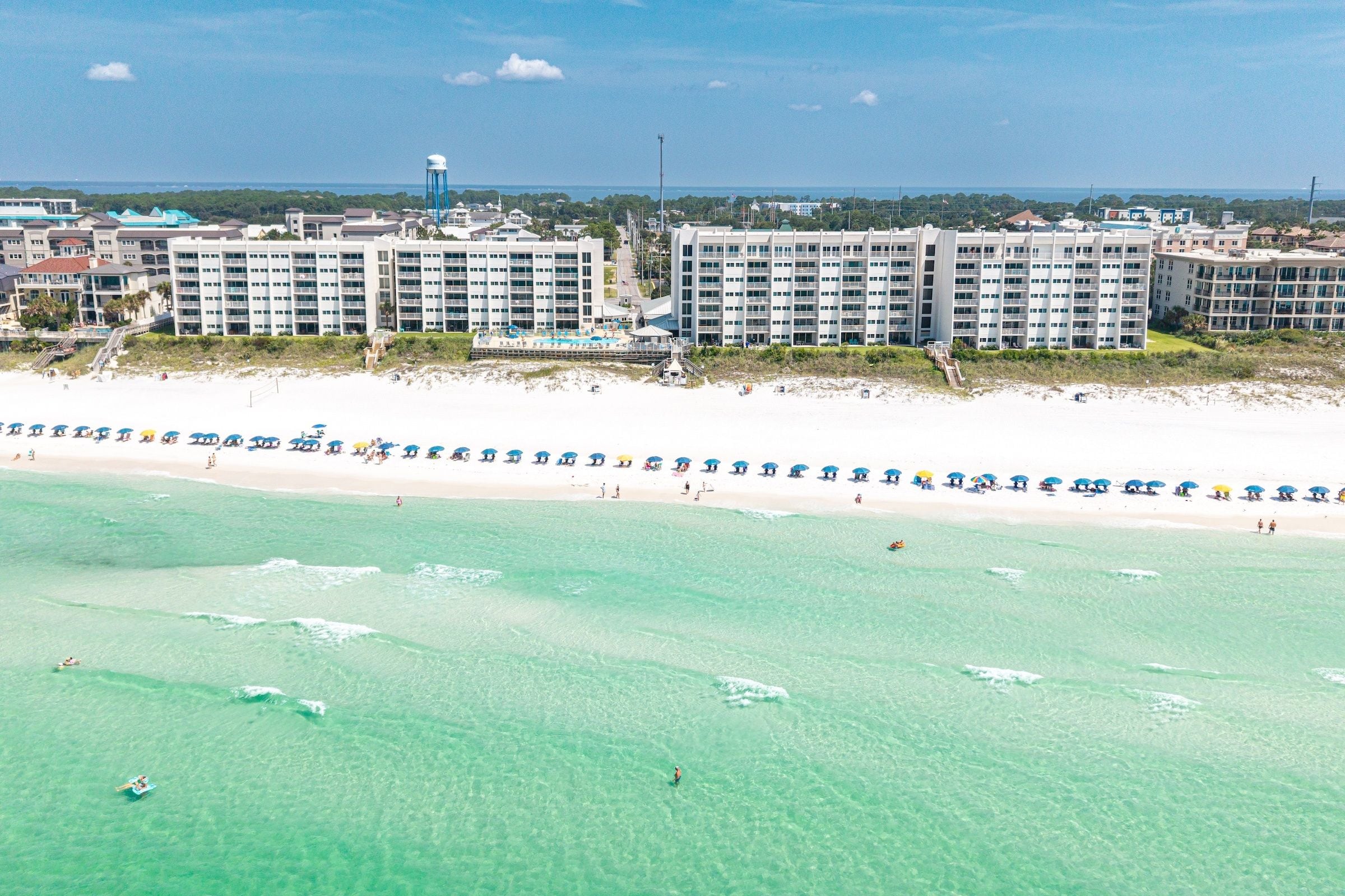Ein Strandhotel mit mehreren Gebäuden, einem Sandstrand und klarem türkisfarbenem Wasser.