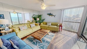A spacious living room with a sofa, coffee table, and a view of the city through large windows. - Mainsail 352 3 Bedroom Condo (Miramar Beach)