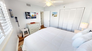 A bedroom with a bed, a television mounted on the wall, a wardrobe, and a small table with a lamp. - Mainsail 352 3 Bedroom Condo (Miramar Beach)