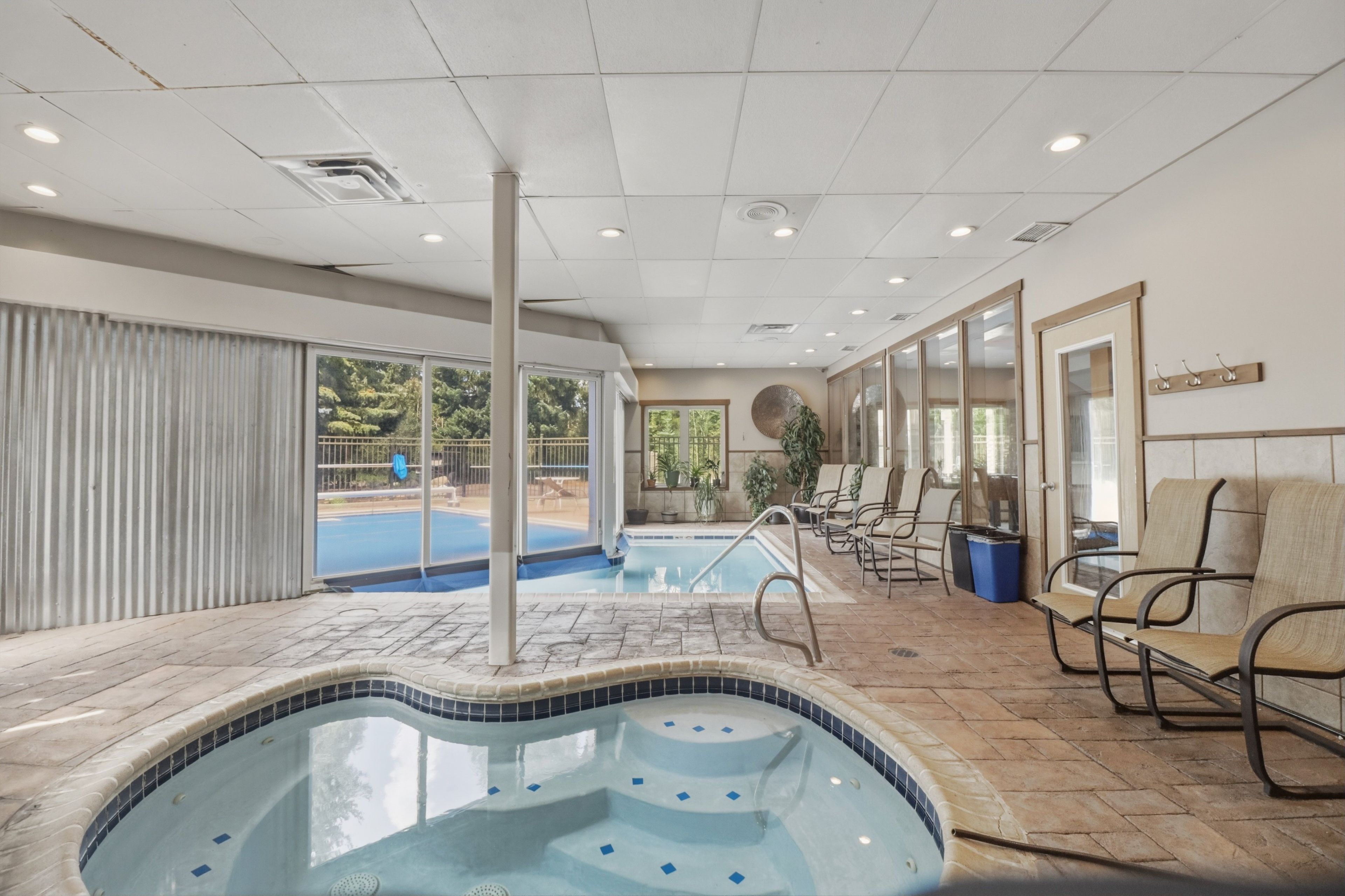 Une piscine intérieure avec des bords carrelés, un espace détente avec des fauteuils et une vue sur l’extérieur grâce à de grandes fenêtres.