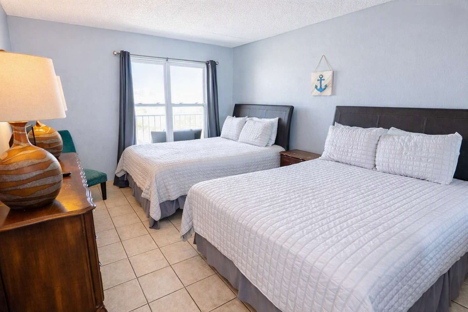 Habitación de hotel con dos camas, una mesita de noche, una lámpara y un ventanal con cortinas.