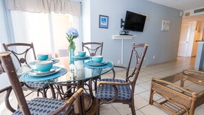 Un coin repas avec une table en verre et des chaises en osier, un téléviseur fixé au mur et une vue sur la pièce adjacente.