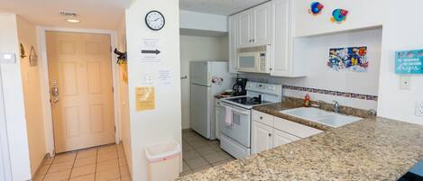 Une cuisine avec des meubles blancs, un plan de travail en granit, un réfrigérateur, un four à micro-ondes et une horloge murale.