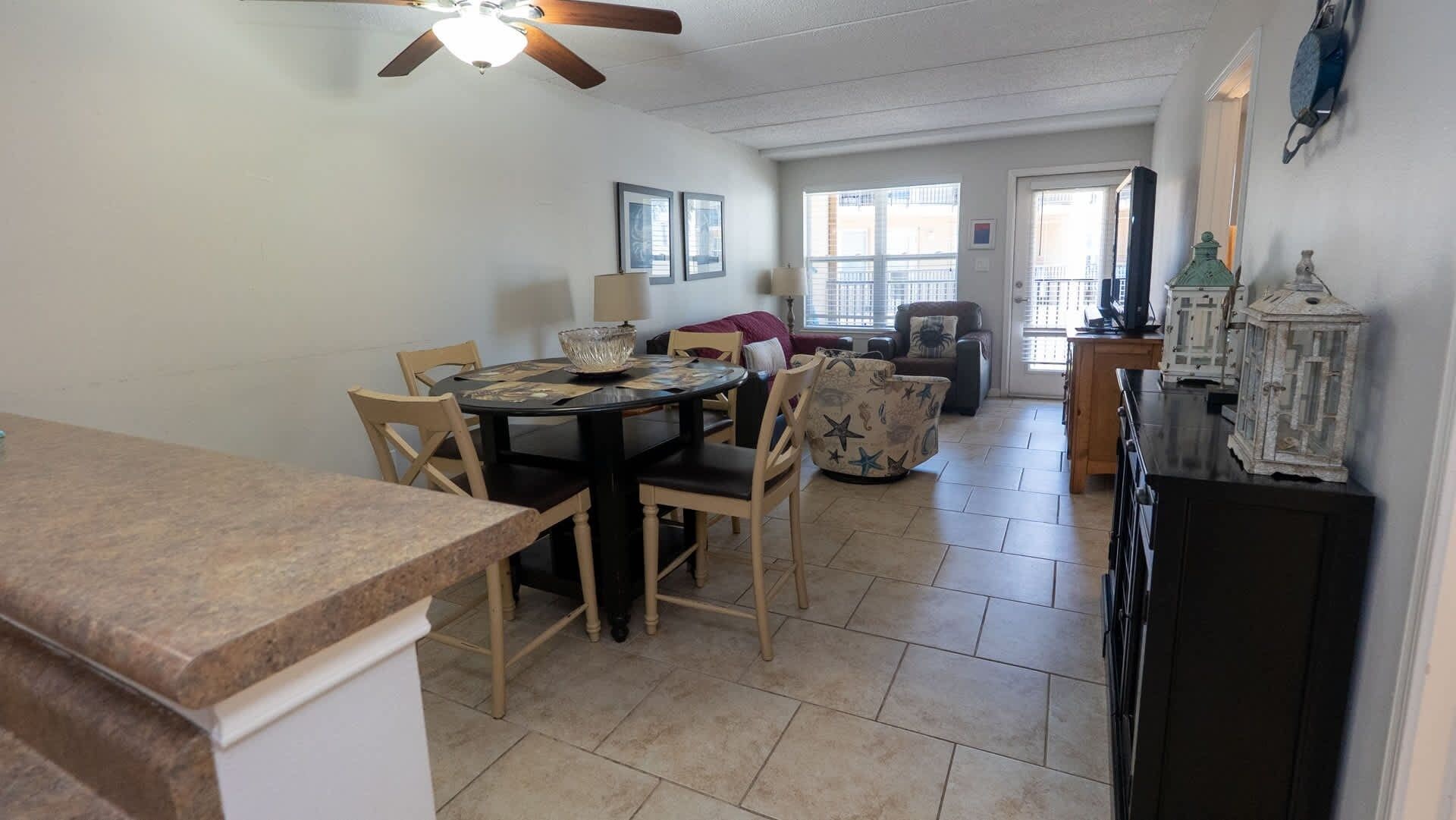 A dining area with a round table, chairs, and a ceiling fan. There is a sofa and a window in the background.