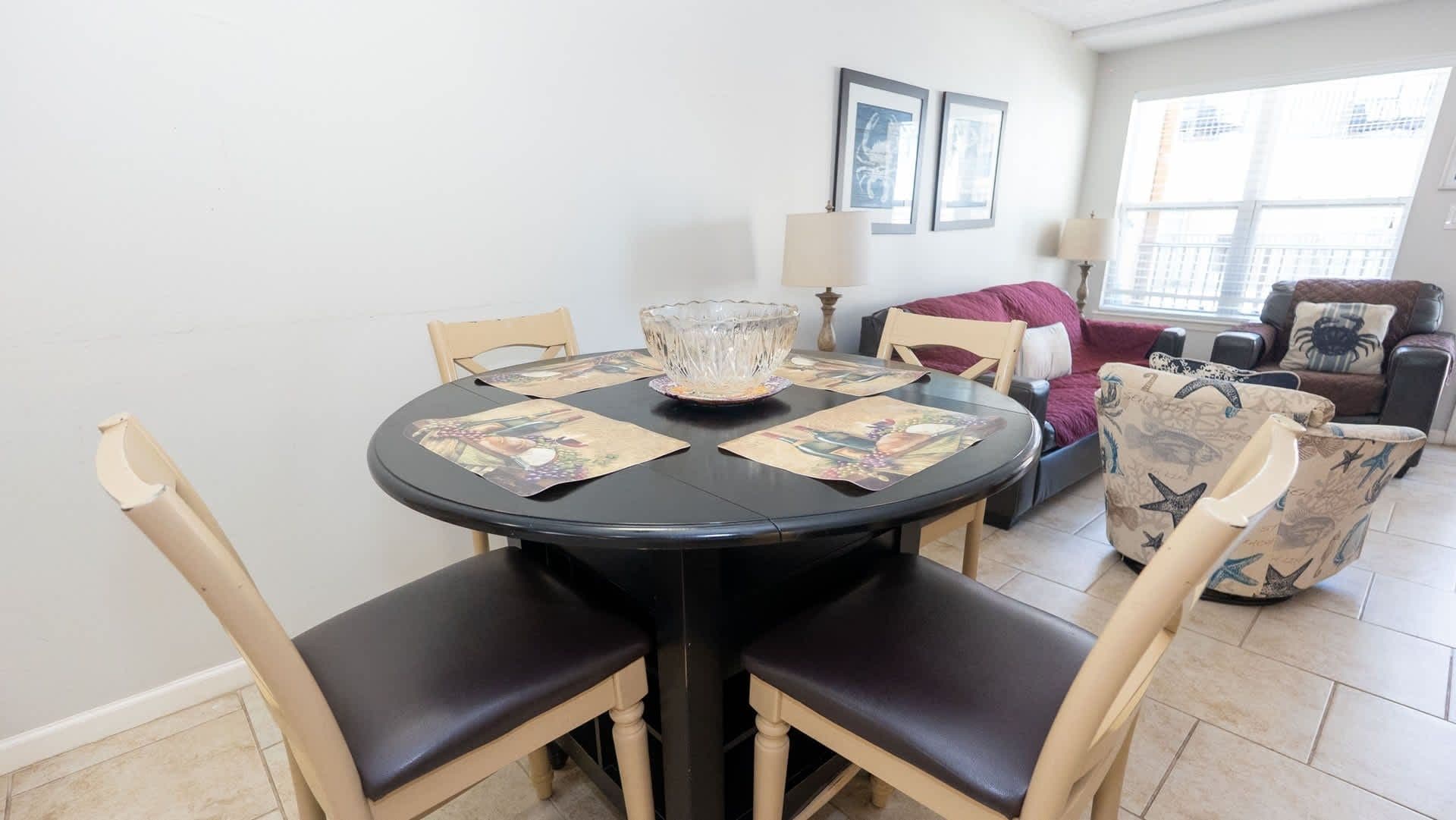 A dining area with a round table, chairs, a glass bowl, and a sofa with a patterned cushion.