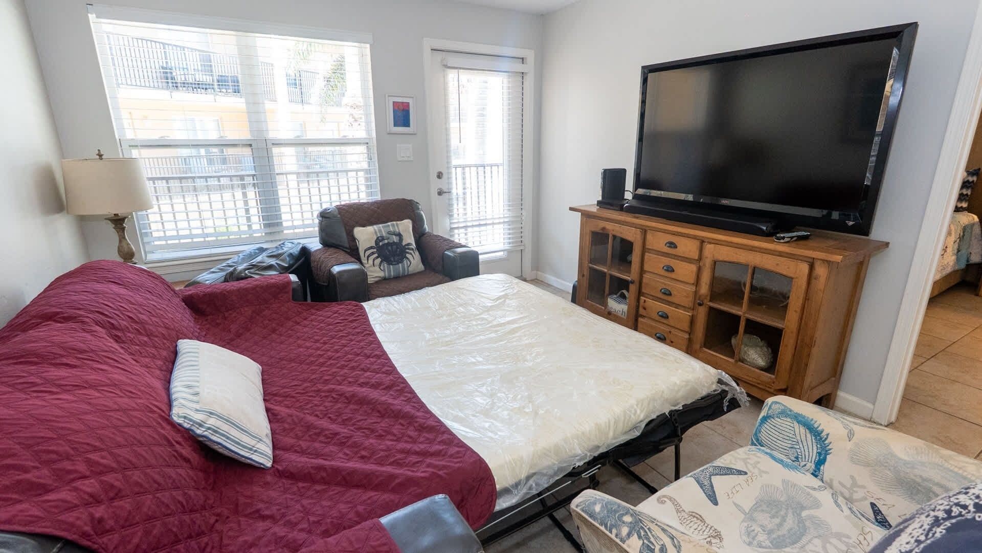 A bedroom with a large bed, a television on a wooden cabinet, a chair, and a window with blinds.